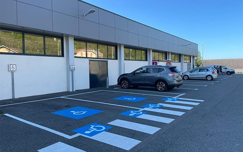 Stationnement aménagé avec marquage au sol pour places PMR devant un bâtiment professionnel, comprenant signalisation bleue et pictogrammes au sol